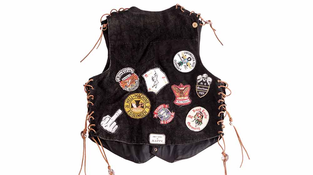 Schwarze Lederweste mit zahlreichen aufgenähten Patches und Aufnähern, darunter Motorrad-Club-Embleme und Aufschriften wie „Hell-Machine“ und „Road Birds MC“. Die Weste hat seitliche Schnürungen und einen kleinen „Don’t worry, be happy“-Aufnäher.