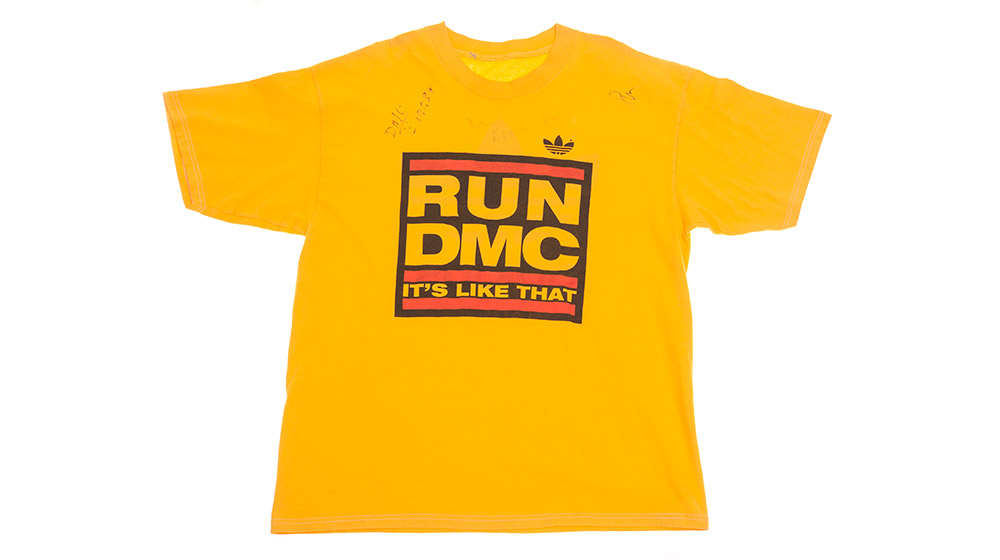 Gelbes T-Shirt mit großem, schwarzem und rot umrandetem Druck „RUN DMC – IT’S LIKE THAT“ und Adidas-Logo auf der Schulter.